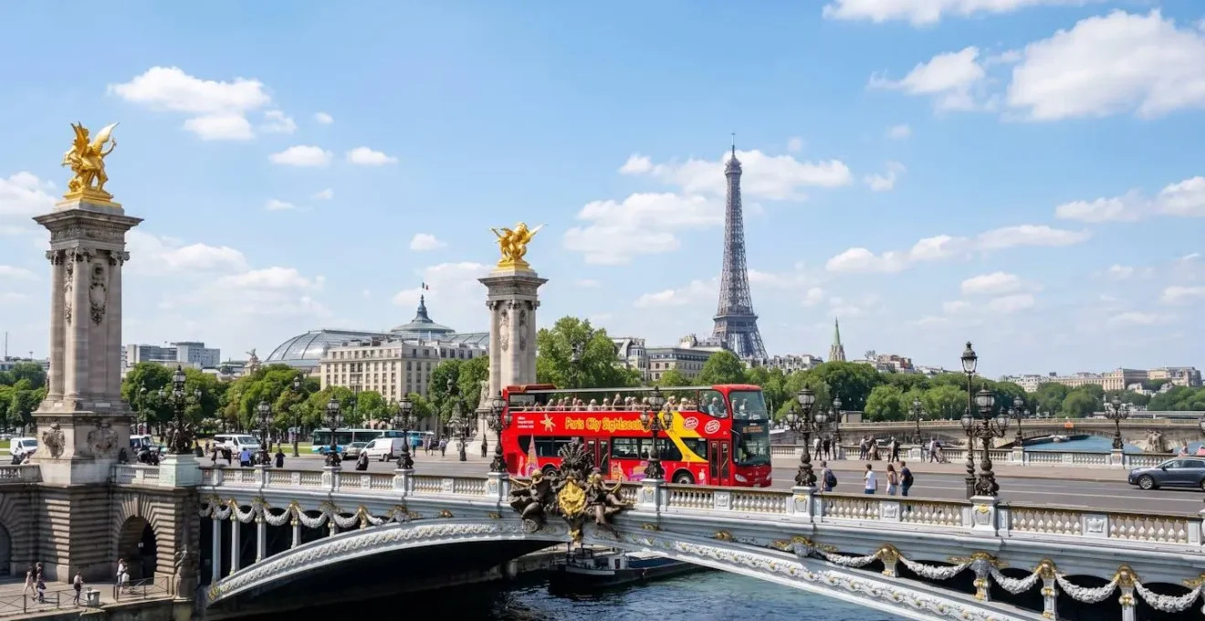 A red open-top tour bus crossing the ornate Pont Alexandre III bridge in Paris on a bright sunny day, with the Eiffel Tower visible in the background and tourists seated on the upper deck