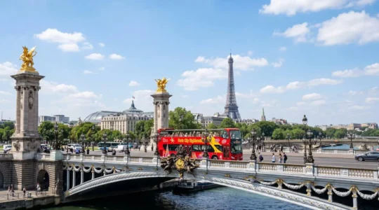 A red open-top tour bus crossing the ornate Pont Alexandre III bridge in Paris on a bright sunny day, with the Eiffel Tower visible in the background and tourists seated on the upper deck
